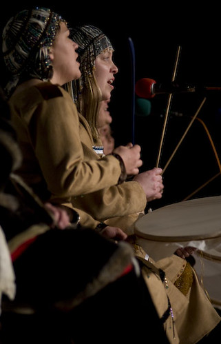 Two young Indigenous women in full regalia play the drums at night