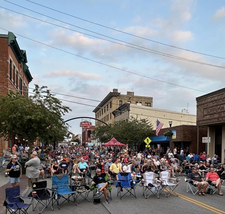 A large crowd is gathered in the middle of a downtown street in Delaware County under sunny skies for a summer concert