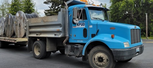 A dump truck carries a flat bed trailer behind it with large spools of fiber loaded onto the back
