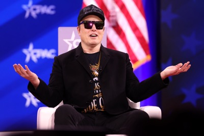 Elon Musk sits on stage at CPAC conference in 2025 wearing sunglasses and MAGA hat