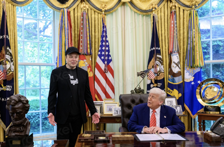 Elon Musk and President Trump in Oval Office together. Trump is seated at desk looking approvingly at Musk who is standing next to desk
