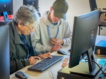 An elderly woman types on a computer keyboard seated next to a youthful instructor in the co-op's Senior Connected program