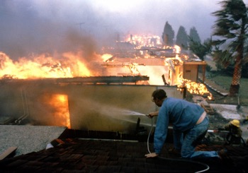 Seven Cabins Fire engulfs home as firefighter kneels on roof next door spraying water with a hose