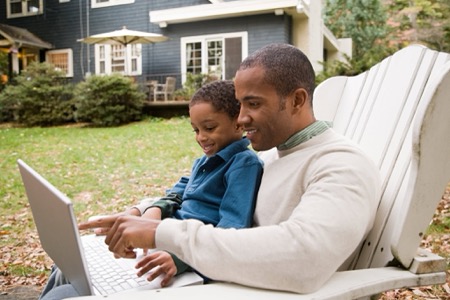 An African-American father and son sit in their front yard looking at a laptop