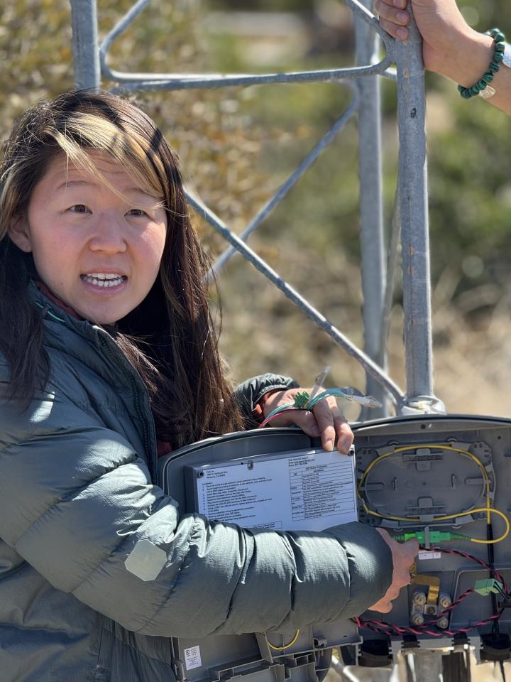 A woman in a green jacket holds open a wireless network access box at the base of a wireless tower