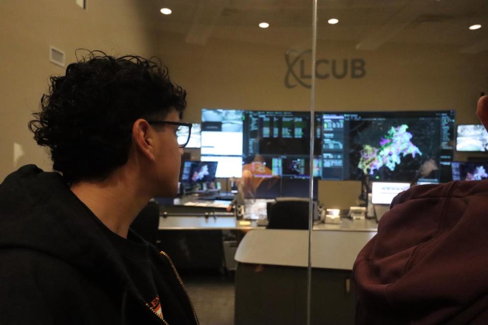 A local high school student on tour of LCUB operations center peers through a window into a control room with monitors arrayed across the back wall