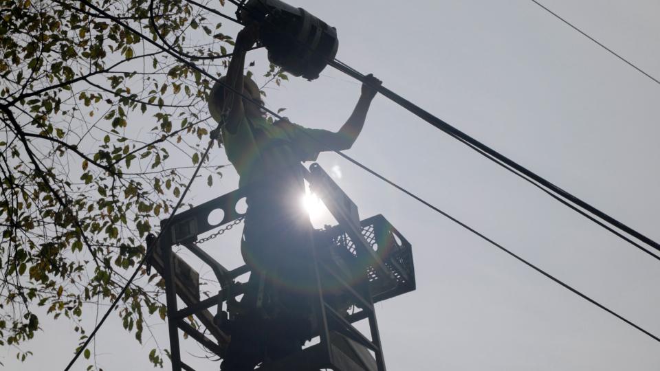 A line work is suspended in air reaching for fiber splice case on aerial fiber line next to a tree