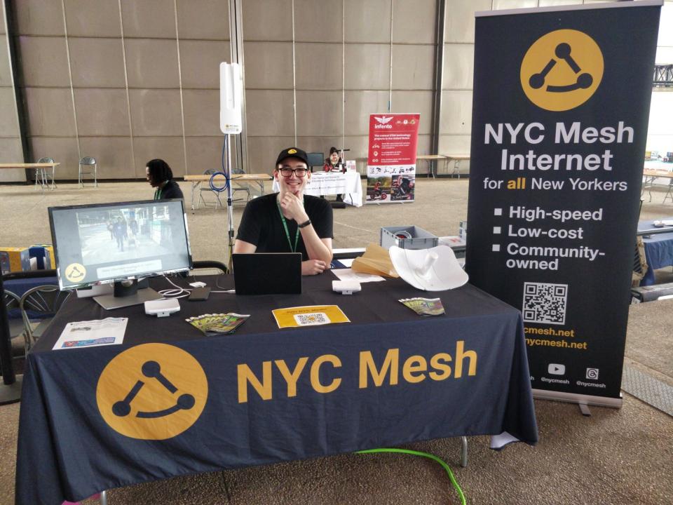 A volunteer sits behind a NYC Mesh table at a Maker Fair in NYC 