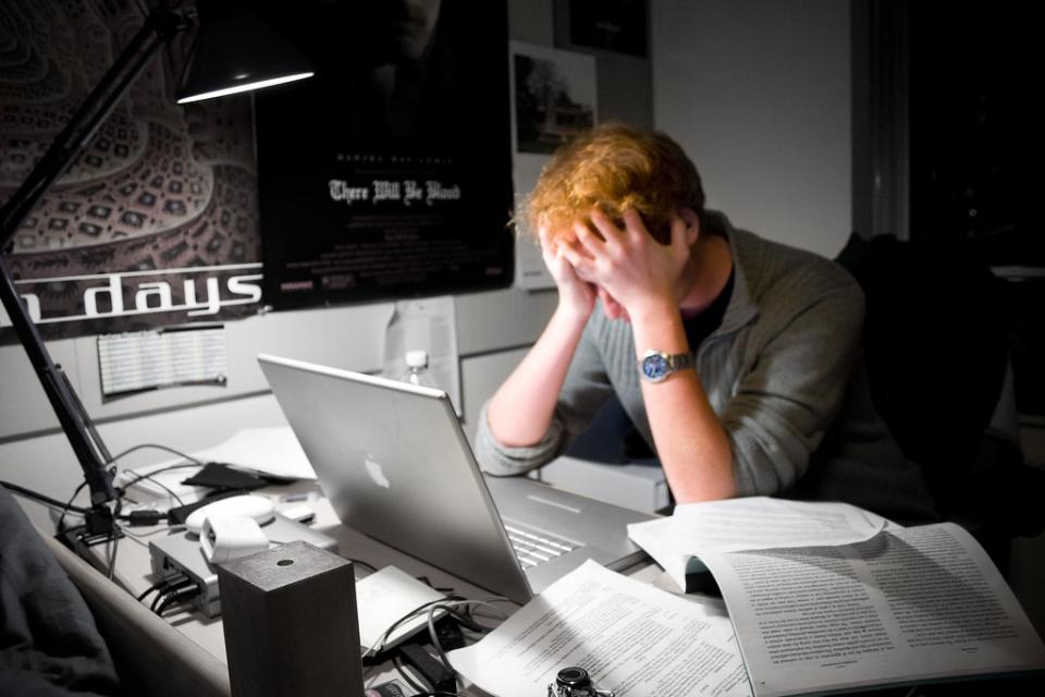 A red-haired man holds his head in his hands in front a computer with paperwork strewn across his desk