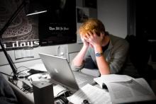 A red-haired man holds his head in his hands in front a computer with paperwork strewn across his desk